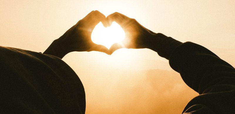 A silhouette of a person's hands forming a heart against a bright sunrise.